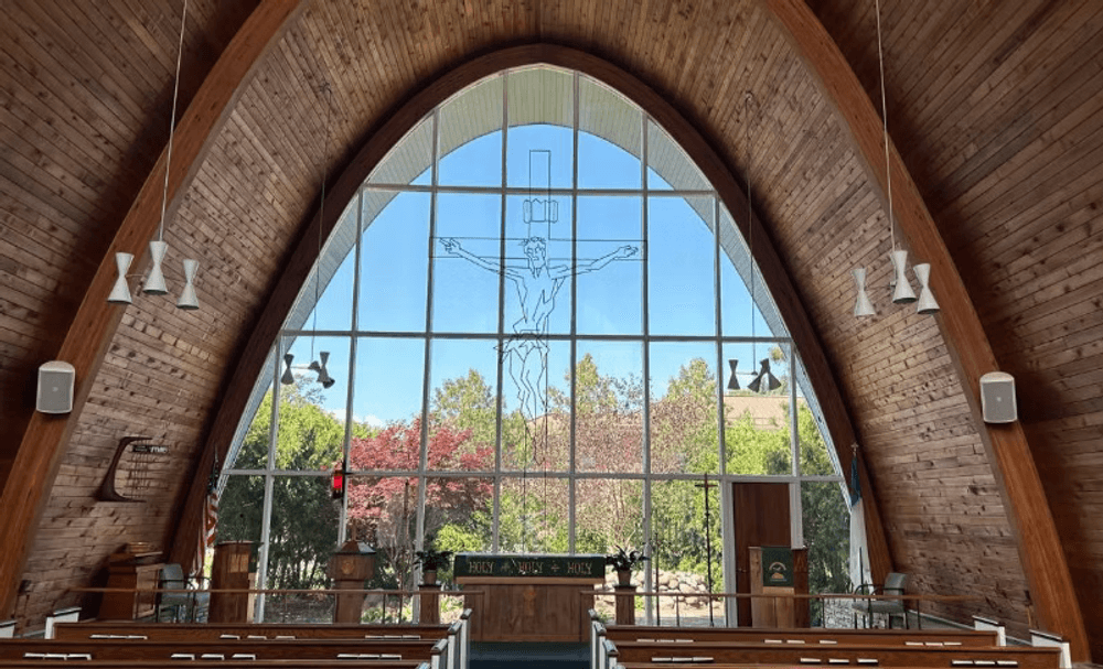 Interior view of a church with a large cross in a wooden arch window and colorful foliage outside.