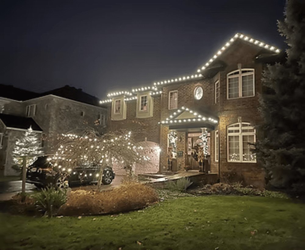 Elegant brick house decorated with holiday lights, surrounded by festive landscaping at night.