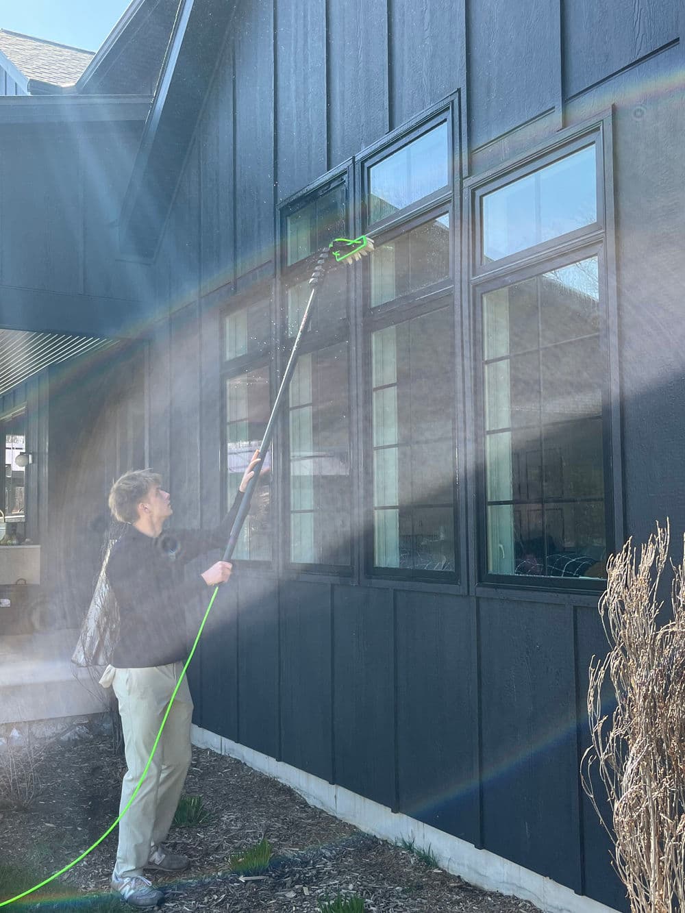 Person cleaning windows on a modern black house with a long pole and water spray.