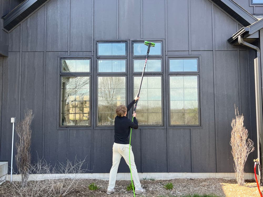 Person cleaning large windows of a modern home using a pole squeegee on a sunny day.