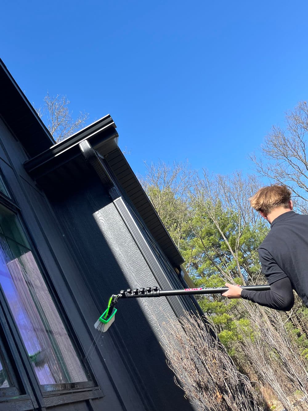 Person using a telescoping pole with brush to clean the exterior of a house under clear blue skies.