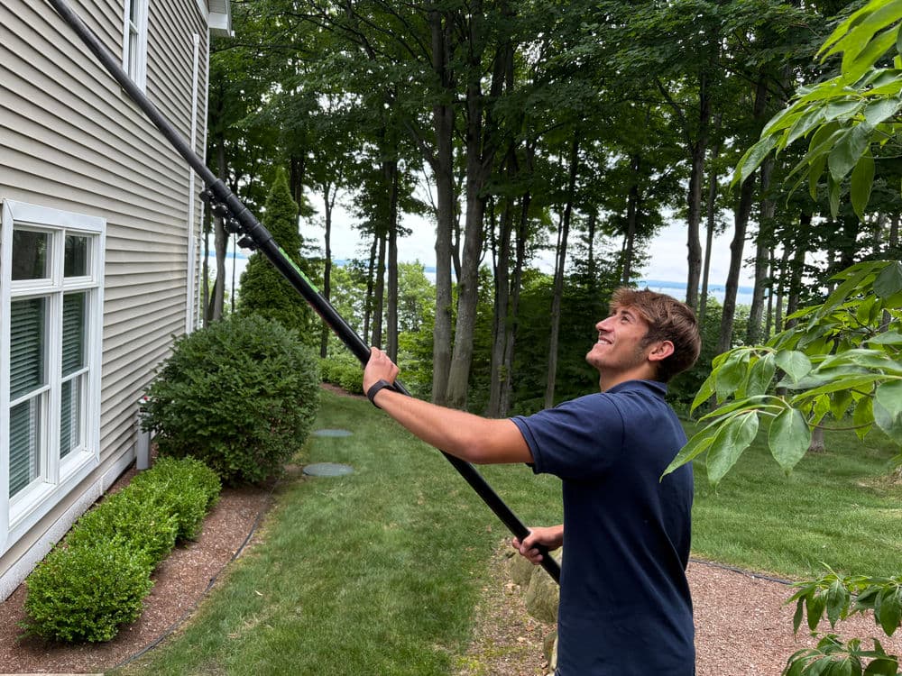 Man using a pole to clean the exterior of a house surrounded by trees and greenery.