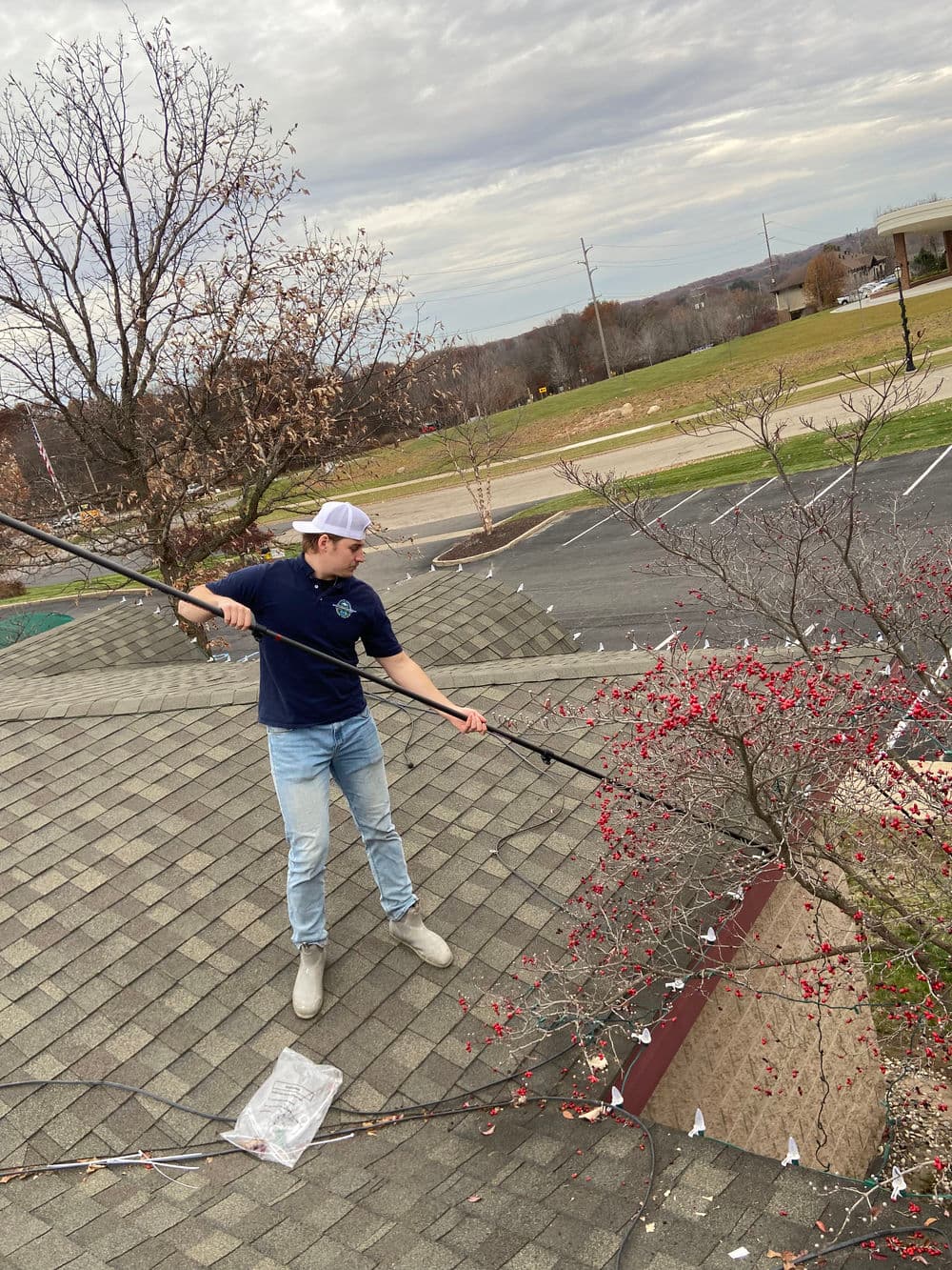 Teen pruning a tree with a pole while standing on a rooftop, cloudy day, holiday lights nearby.
