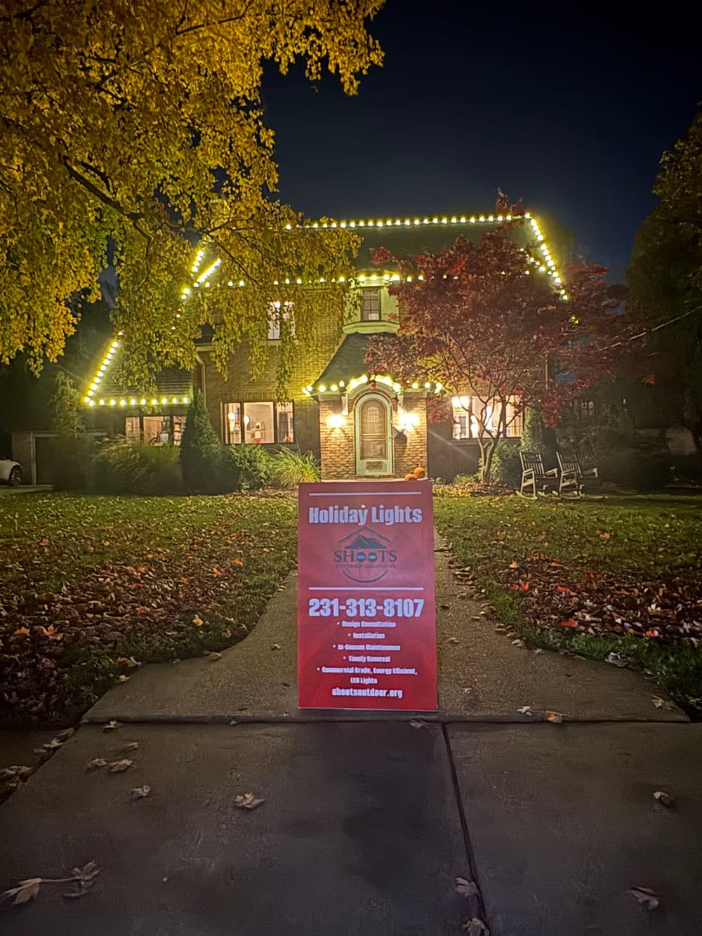 Holiday lights display on a house with a promotional sign for Shoots Holiday Lights.