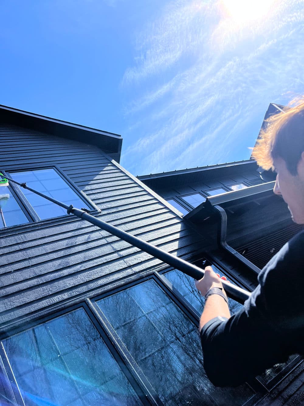 Person using a pole to clean windows of a modern black house under a bright blue sky.