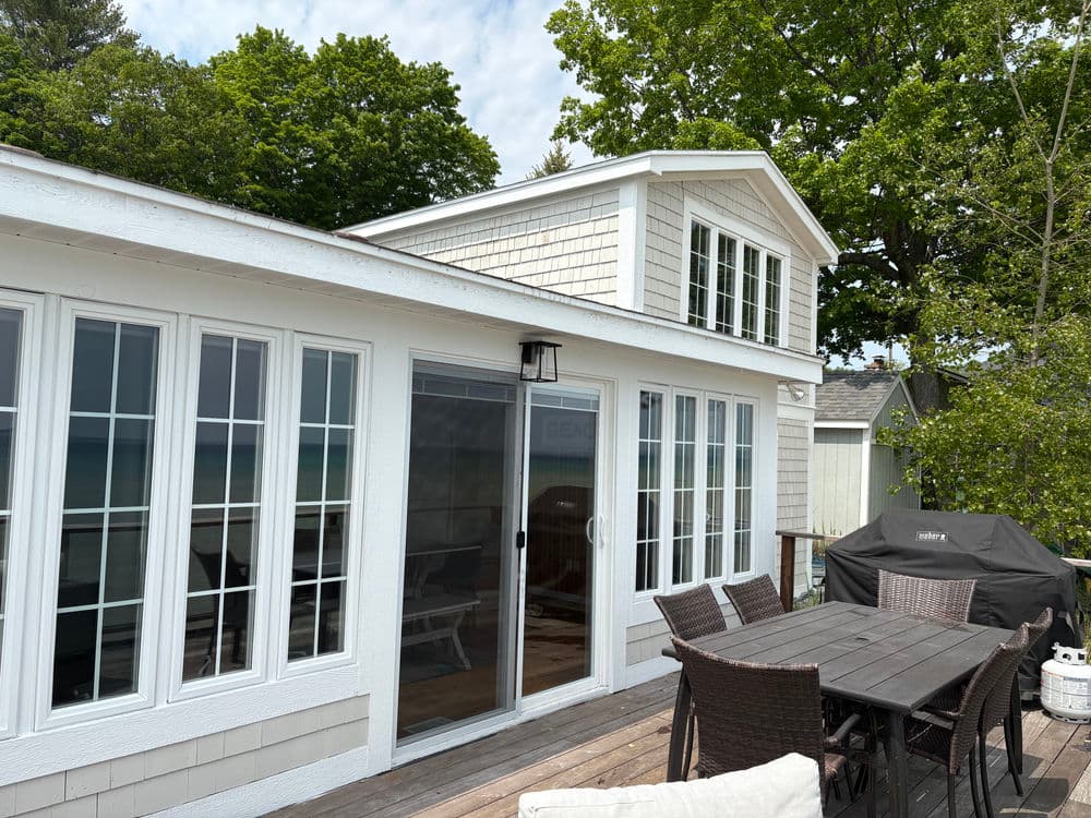 Cozy lakeside house with large windows, outdoor dining area, and grill surrounded by trees.