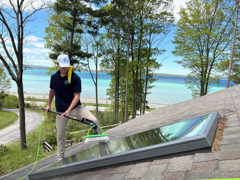 Person cleaning a skylight on a sloped roof with a lake view and trees in the background.