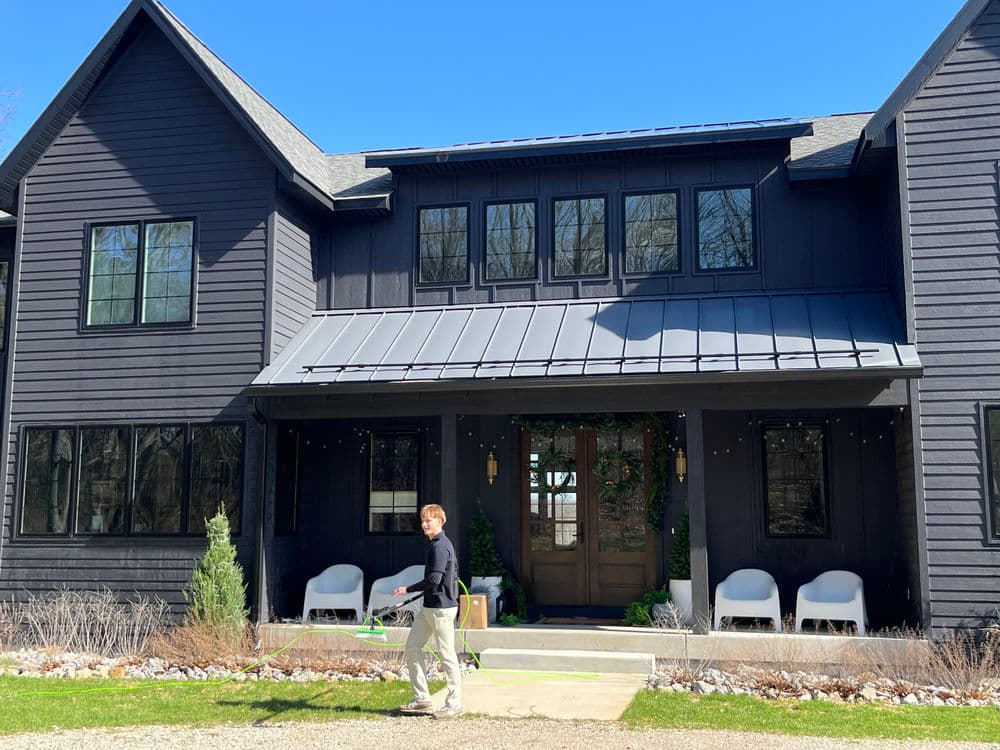 Man walking with a lawn mower in front of a modern black house with large windows.