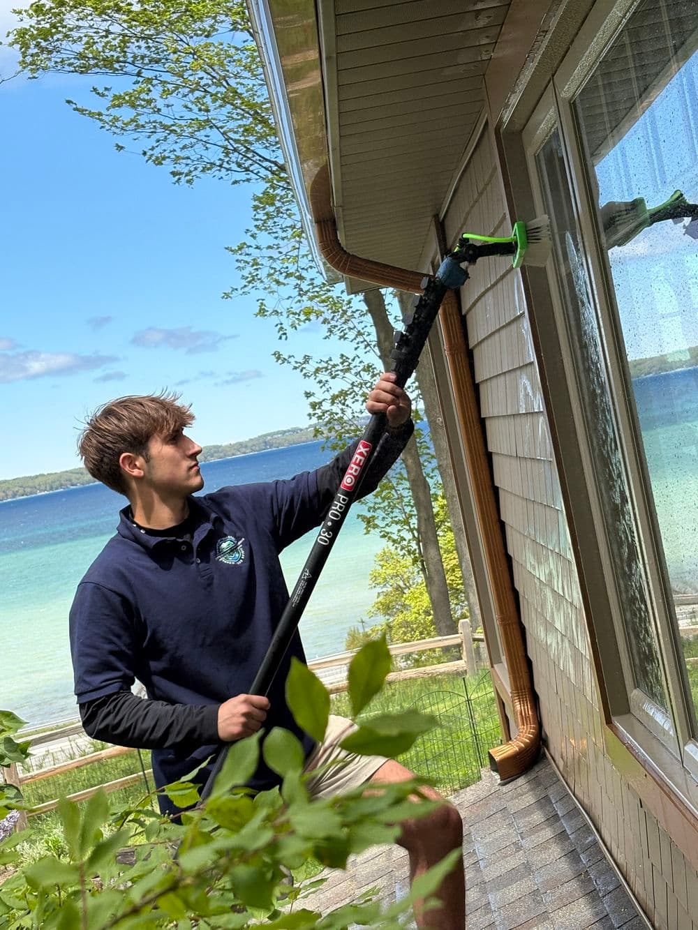 Person using a long pole to clean a window on a house near a lake with trees in the background.